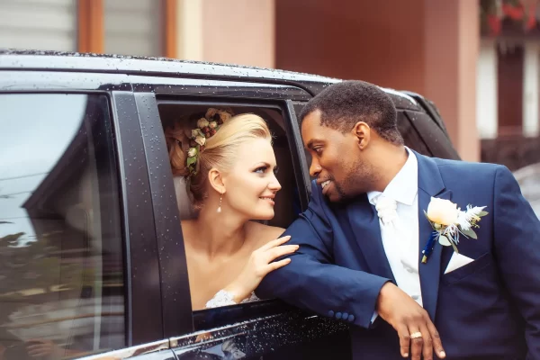 Happy newlyweds of beautiful bride woman in car and african American groom man outdoors on rainy wedding day